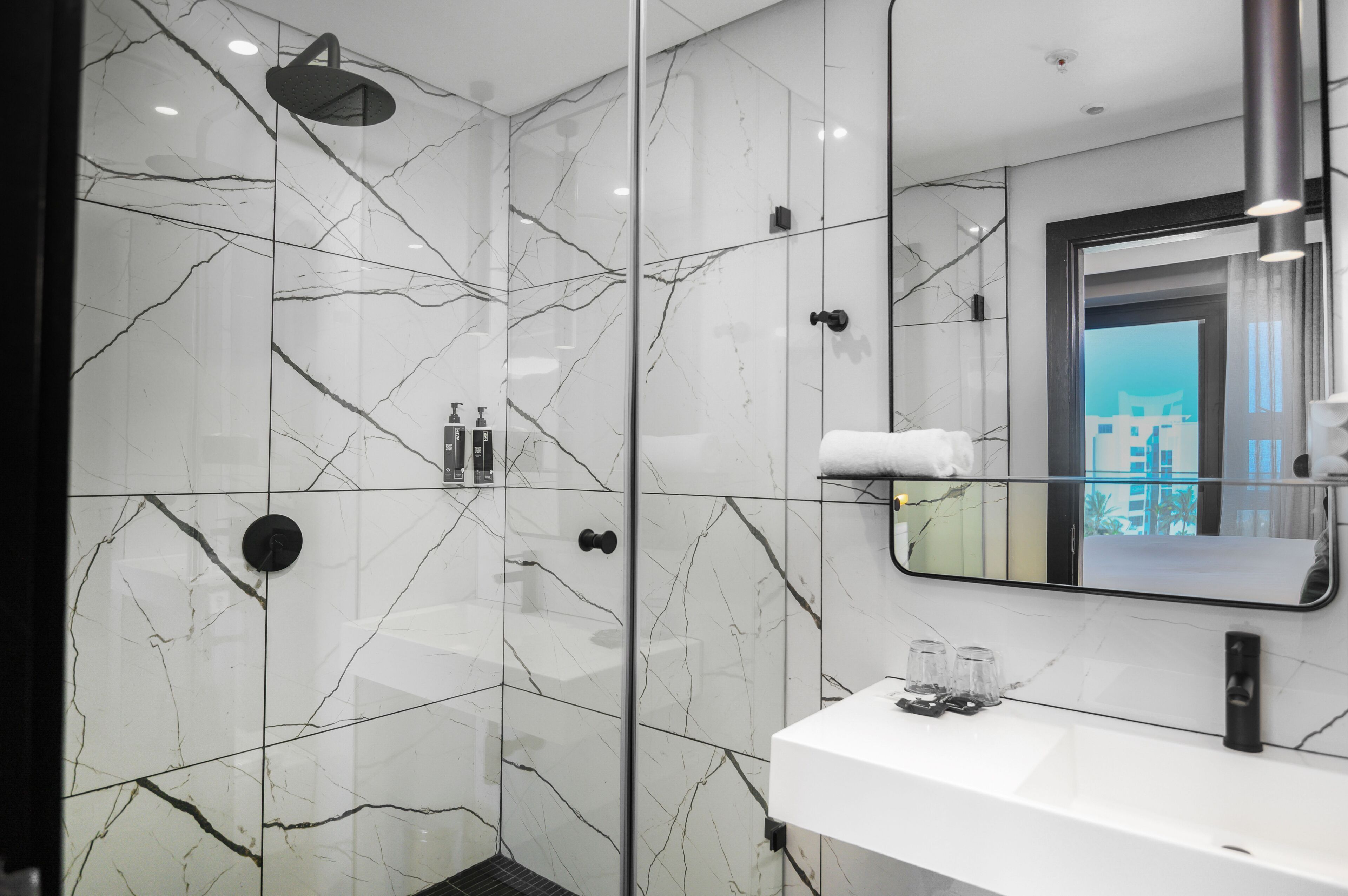 A modern bathroom with a glass-enclosed shower, a white marble backsplash, and a large mirror reflecting a cityscape.