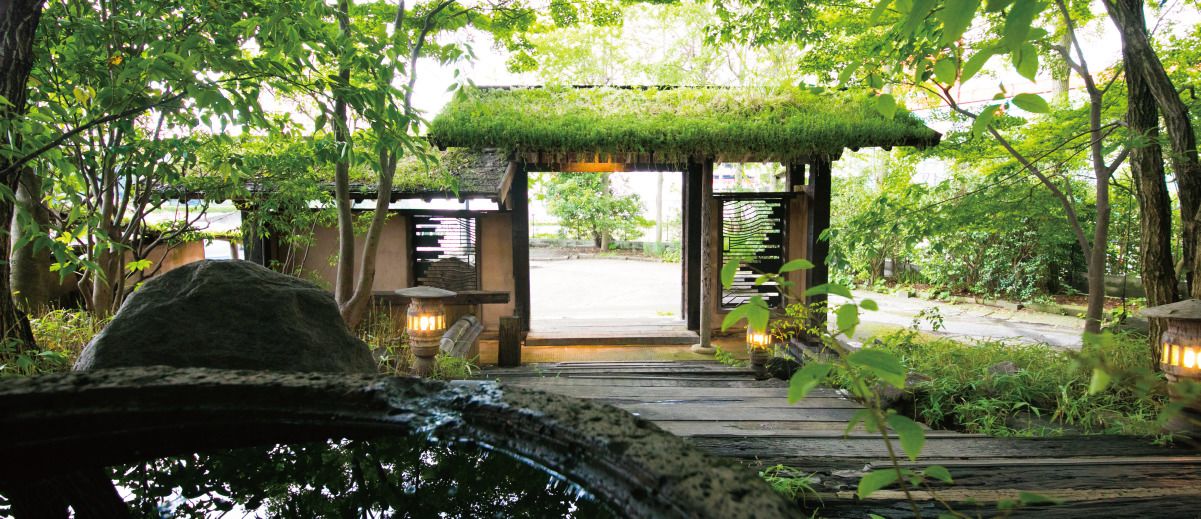 A wooden pathway leading to a traditional Japanese-style entrance with a thatched roof, lanterns, and a stone lantern.