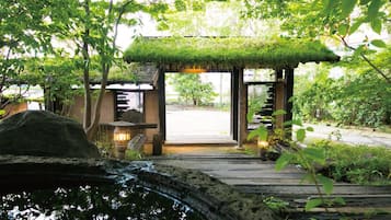 A wooden pathway leading to a traditional Japanese-style entrance with a thatched roof, lanterns, and a stone lantern.