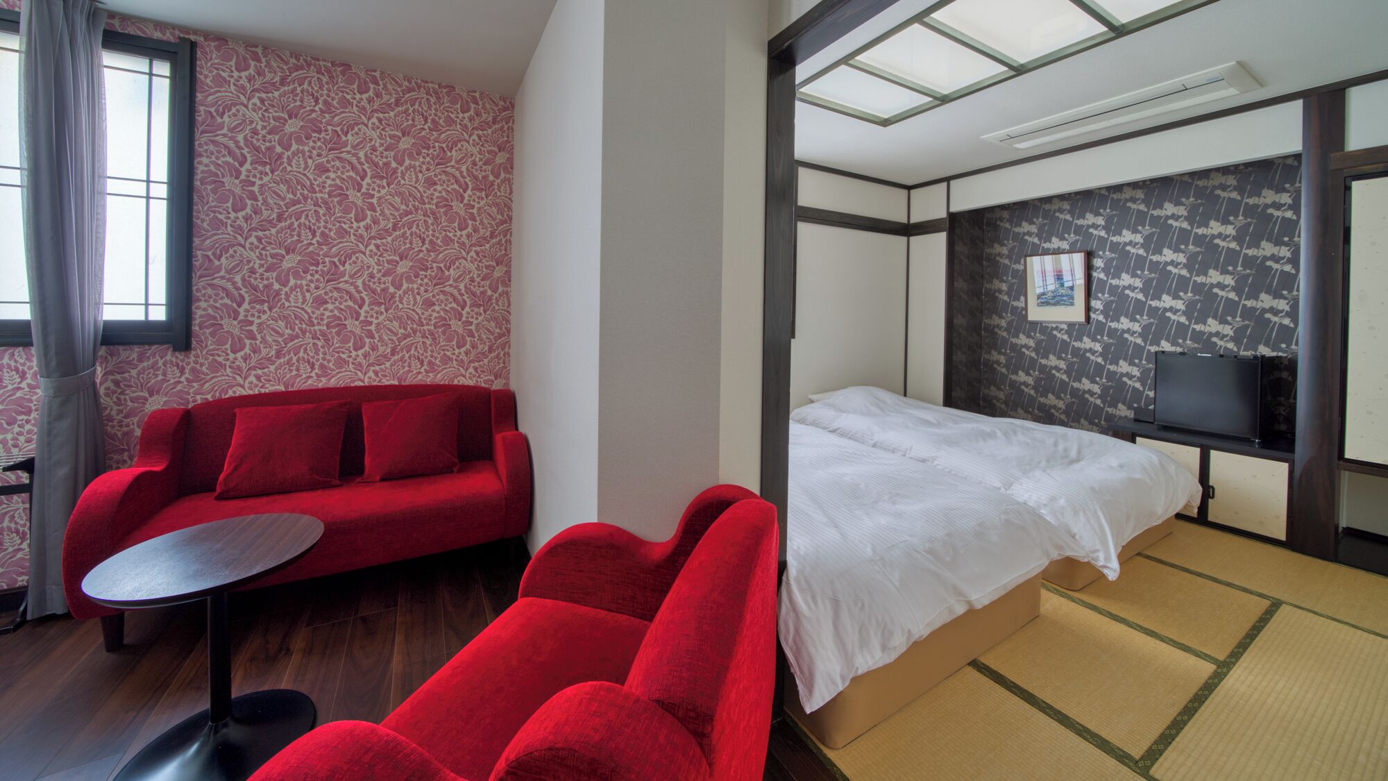 Une chambre d’hôtel avec un canapé rouge, une petite table ronde, un lit avec une literie blanche, un téléviseur posé sur un meuble et un mur à motifs.