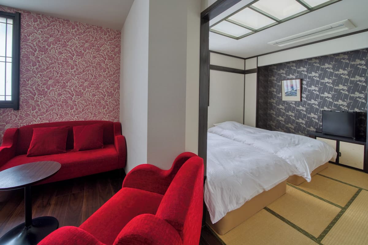 Une chambre d’hôtel avec un canapé rouge, une petite table ronde, un lit avec une literie blanche, un téléviseur posé sur un meuble et un mur à motifs.