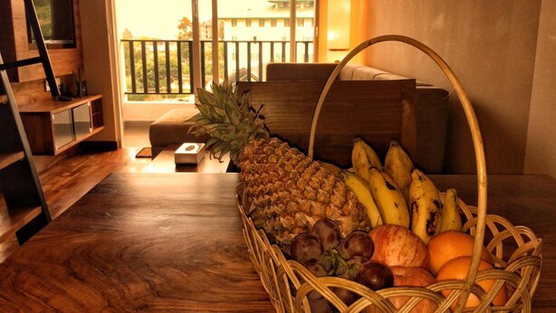 Un panier en osier rempli de fruits est posé sur une table en bois dans un salon donnant sur un balcon.