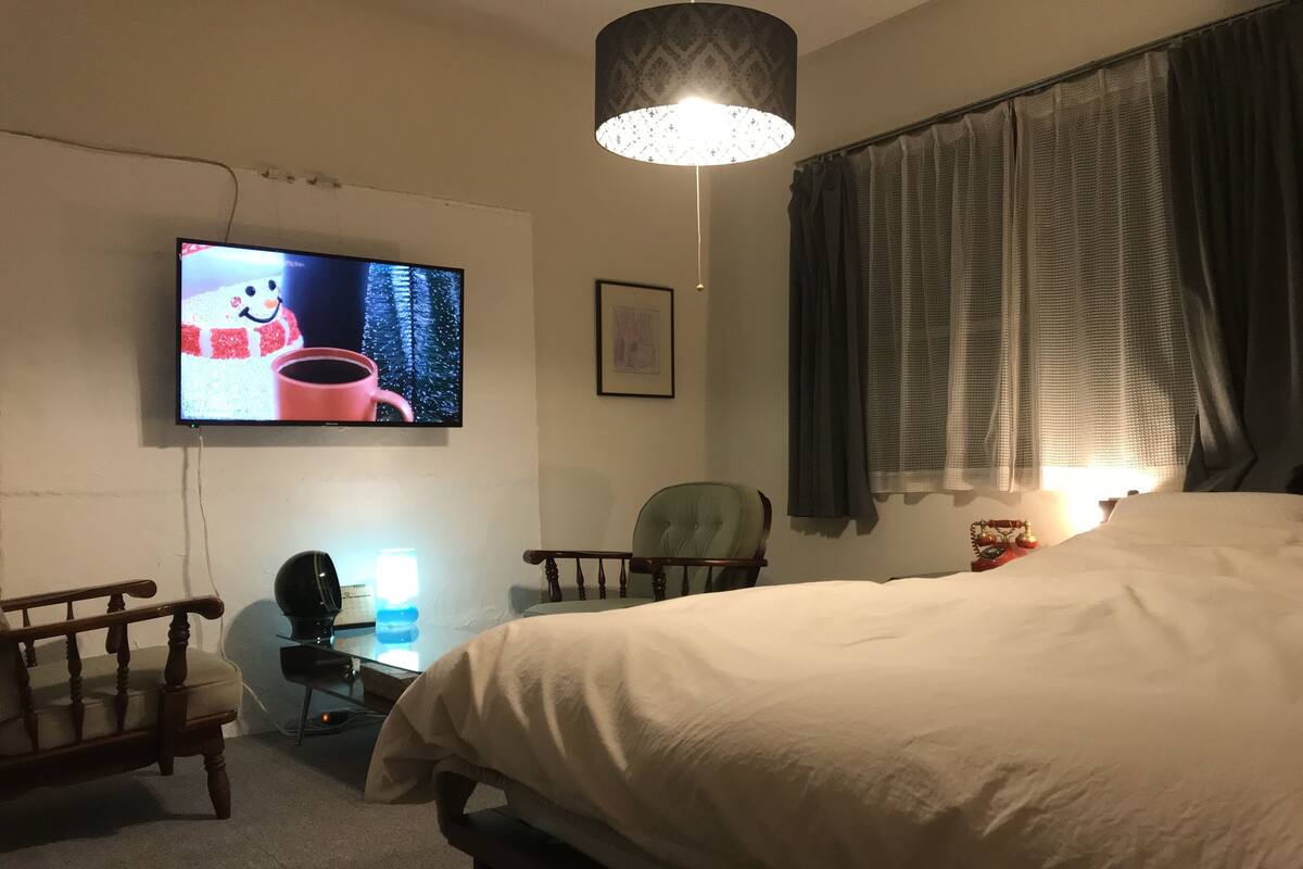 A bedroom with a large bed, a television mounted on the wall, a desk with a lamp, and a chair.