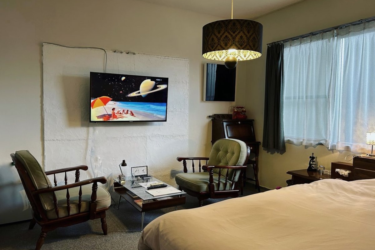 A hotel room with a bed, two armchairs, a desk with a lamp, a television on the wall, and a window with curtains.