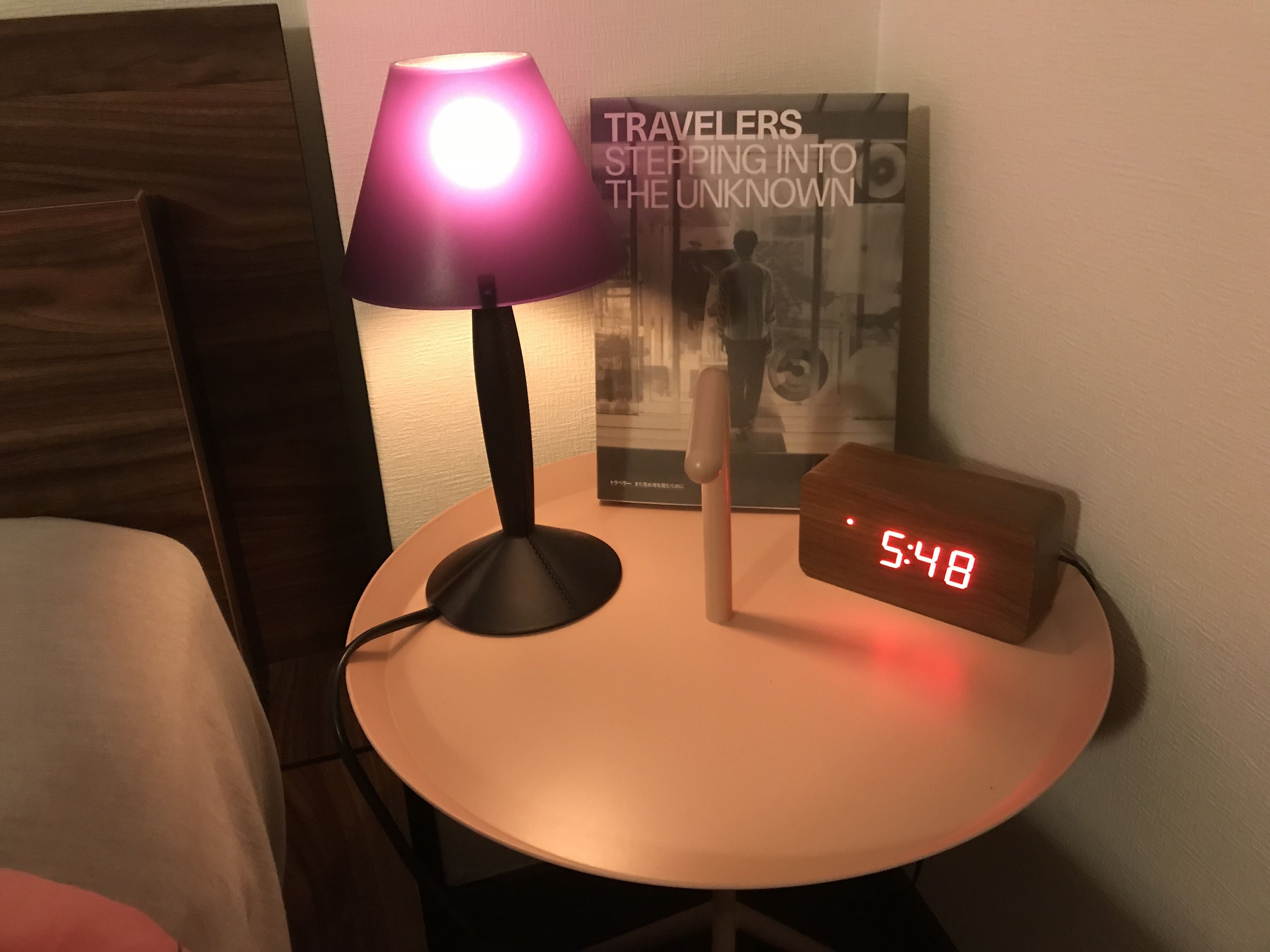 Uma pequena mesa redonda com um relógio digital mostrando 5:48, uma luminária roxa e uma moldura com a foto intitulada 'VIAJANTES DESCENDENDO AO DESCONHECIDO'.
