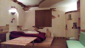 Una habitación con una cama, una mesa y un banco. Hay una ventana con cortina, una lámpara de pared y un armario de madera.