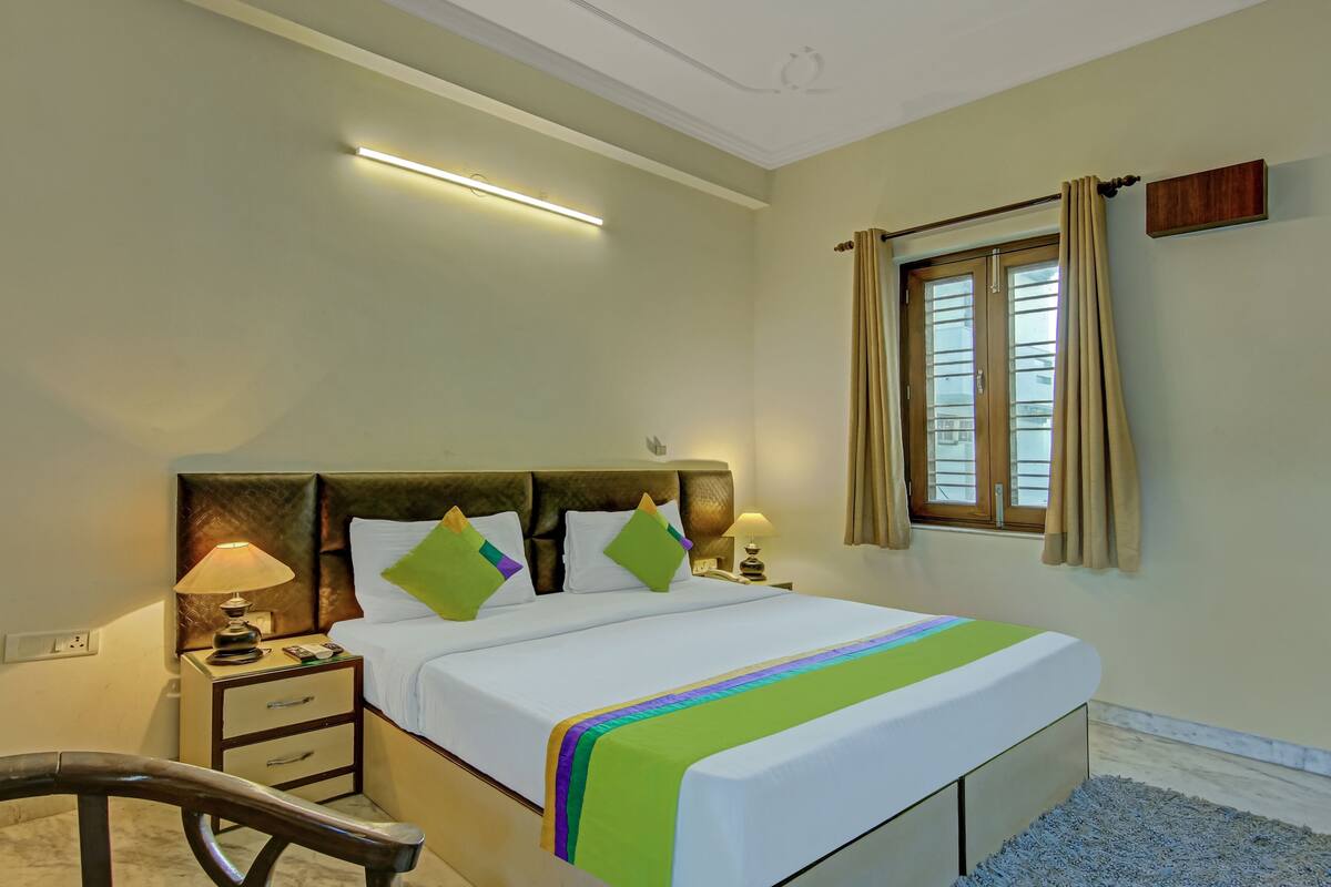 A hotel room with a bed, bedside tables, a lamp, a chair, a window with curtains, and a ceiling light.