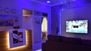 Un’area reception con una scrivania in legno che mostra un logo, uno schermo televisivo, un divano e una parete con quadri appesi.