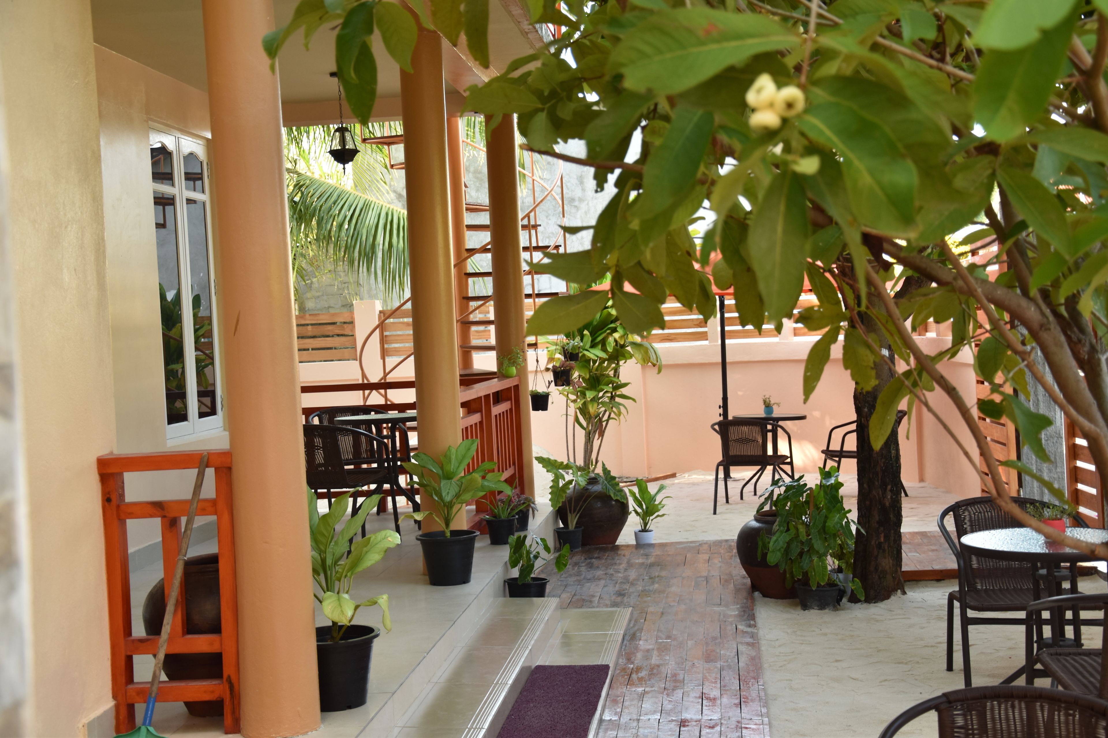 A covered outdoor seating area with potted plants, wooden furniture, and a tiled floor.