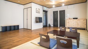 A spacious room with a wooden floor, a large table with chairs, a bench, and a cabinet with drawers. - Guesthouse Kyozen (Kyoto)