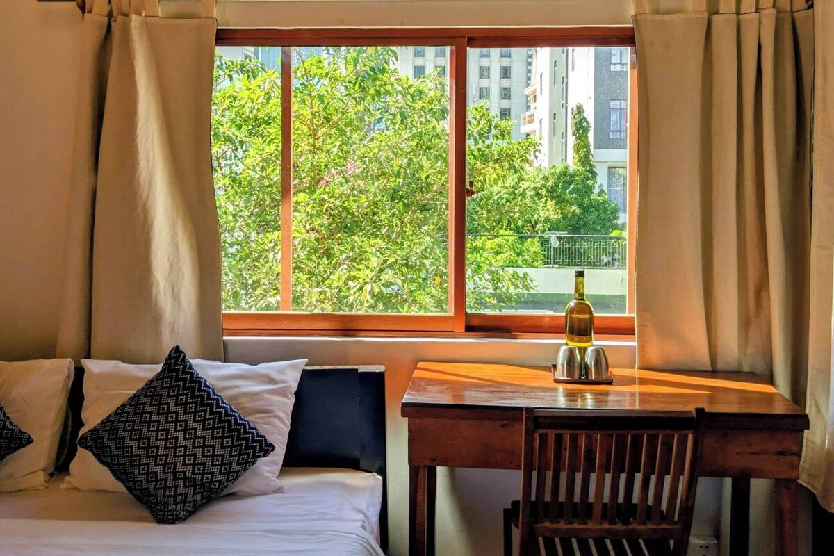 A hotel room with a bed, a desk, and a chair. There is a window with curtains, a wall-mounted air conditioner, and a lamp.