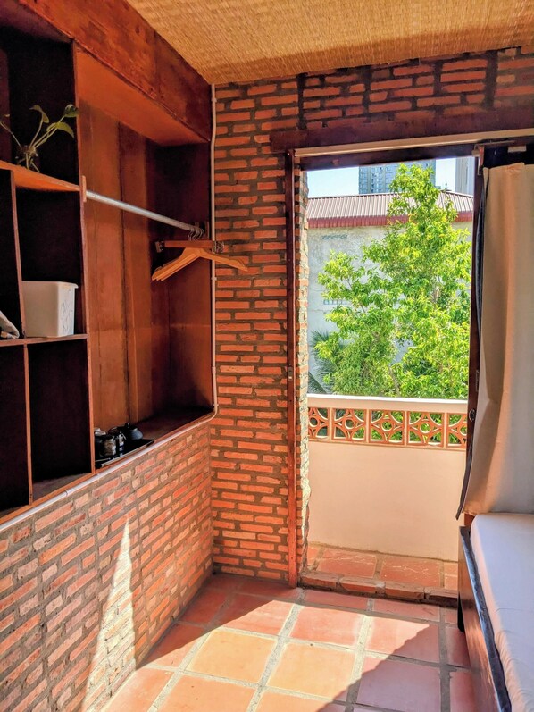 Un balcón con un muro de ladrillo, un estante de madera con frascos, y vistas a un edificio y árboles.