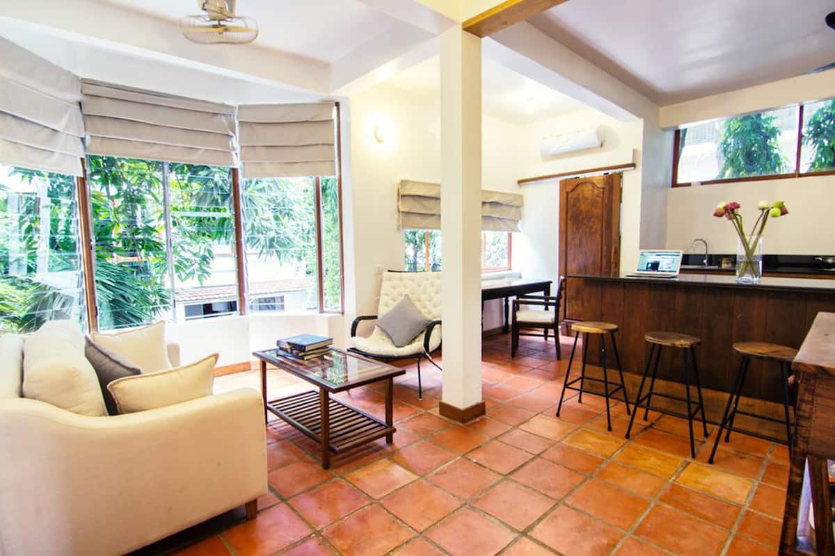 A spacious living area with a sofa, coffee table, and bar stools. Large windows offer a view of greenery outside.