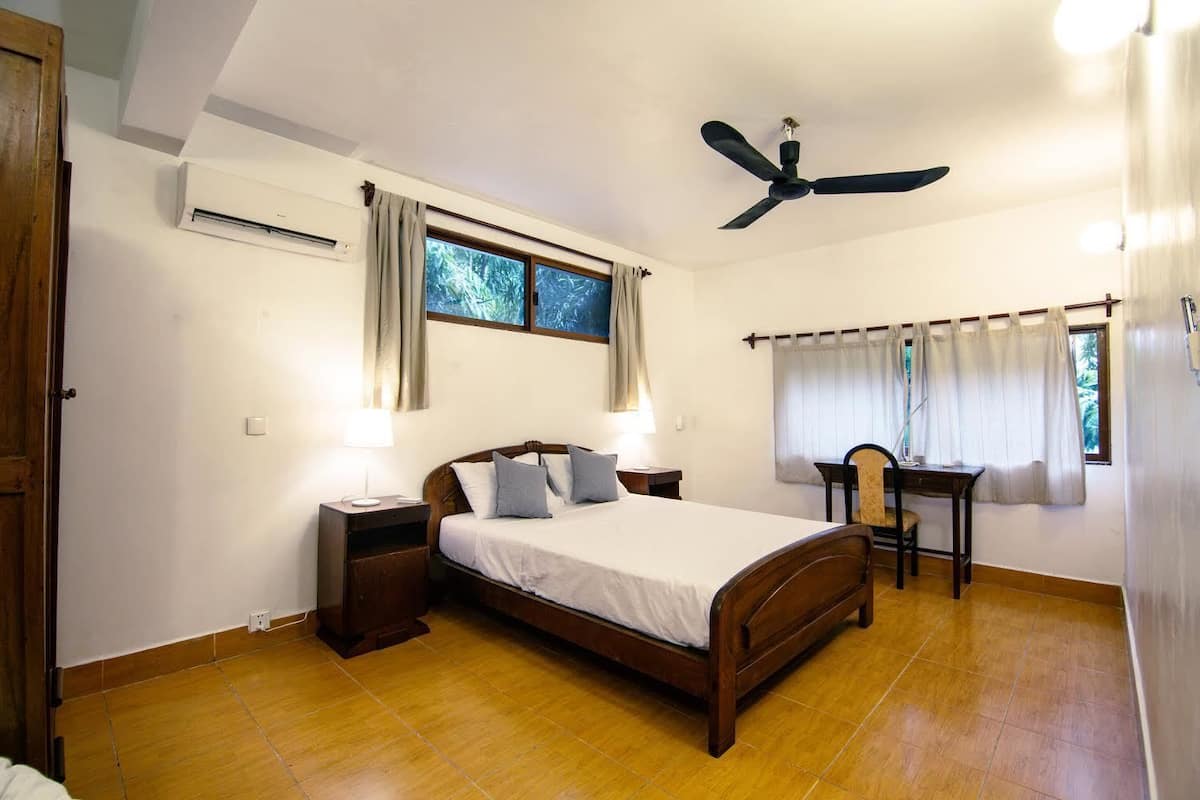 A bedroom with a bed, bedside tables, a desk, a chair, a ceiling fan, and a window with curtains.
