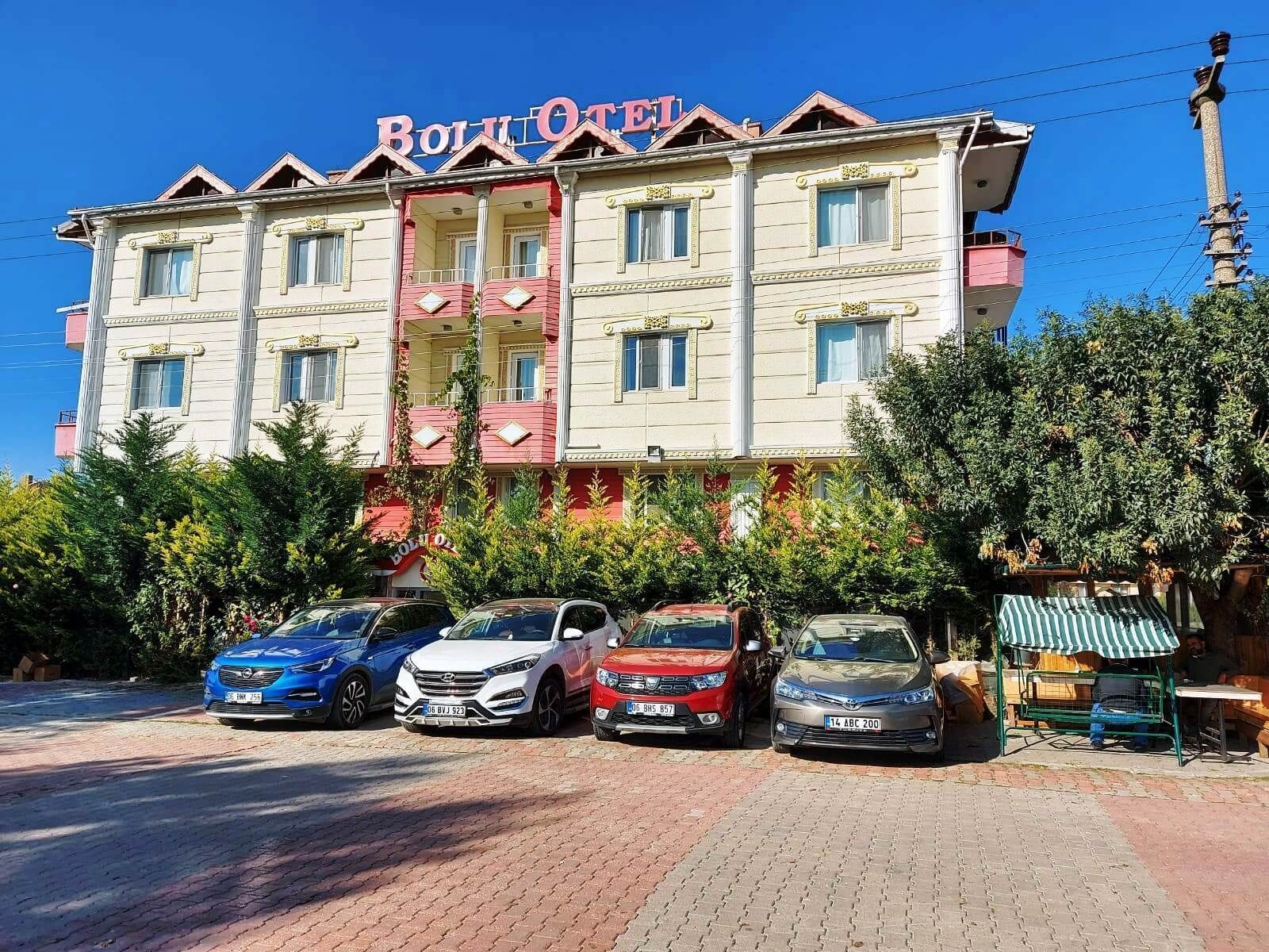 A hotel with a red and white facade, multiple balconies, and a parking area with several cars.