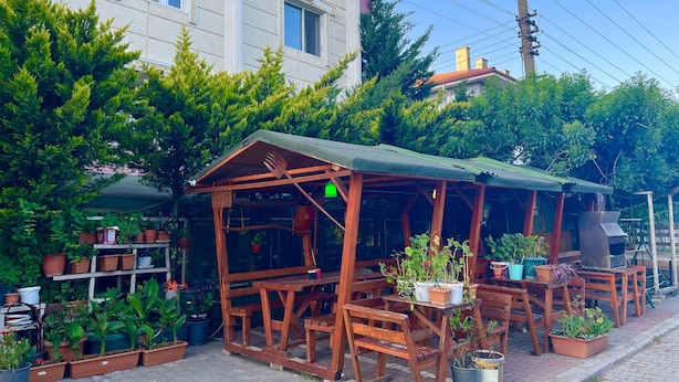 Un espace extérieur aménagé avec des meubles en bois, comprenant des tables et des chaises, entouré de plantes en pot et avec un bâtiment aux grandes fenêtres en arrière-plan.