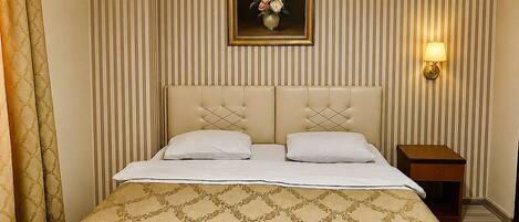 A hotel room with a large bed, a nightstand, a wall-mounted lamp, and a framed floral painting.