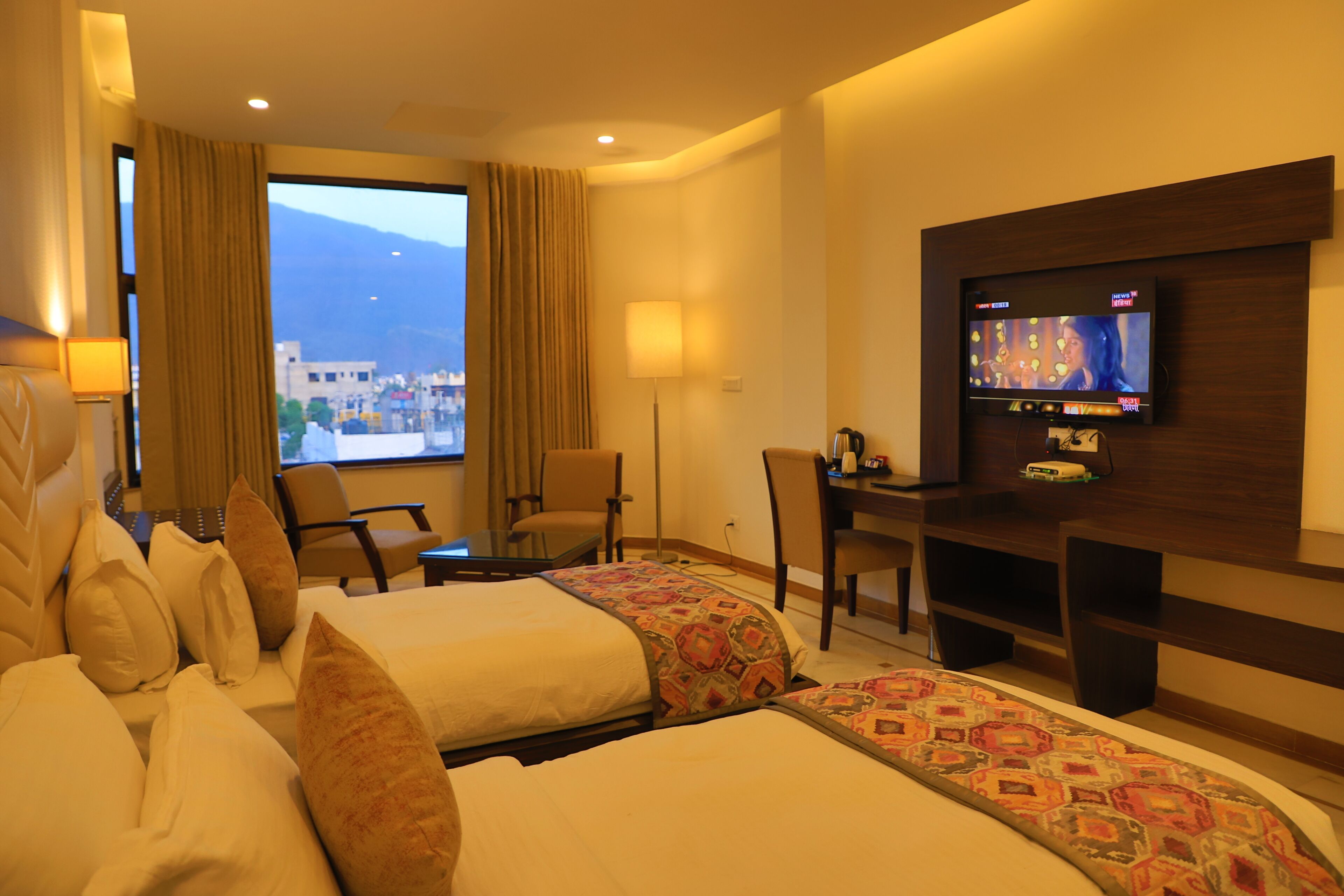 A hotel room with two beds, a desk, a TV, and a large window with a mountain view.