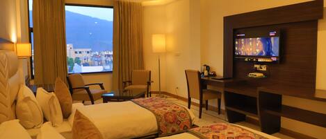 A hotel room with two beds, a desk, a TV, and a large window with a mountain view.