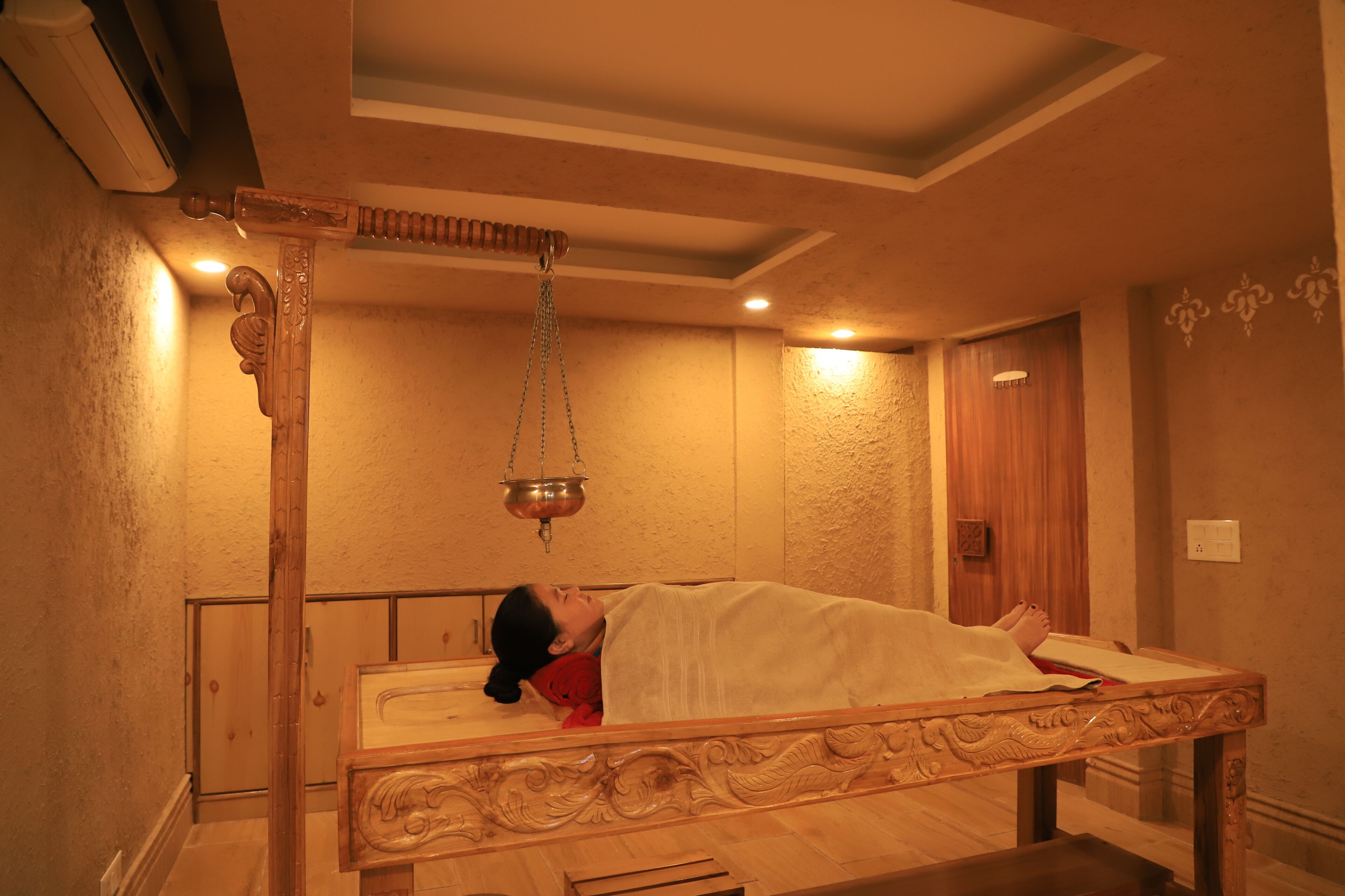 A traditional wooden bed with intricate carvings, a hanging copper vessel, and a wooden door in a warmly lit room.