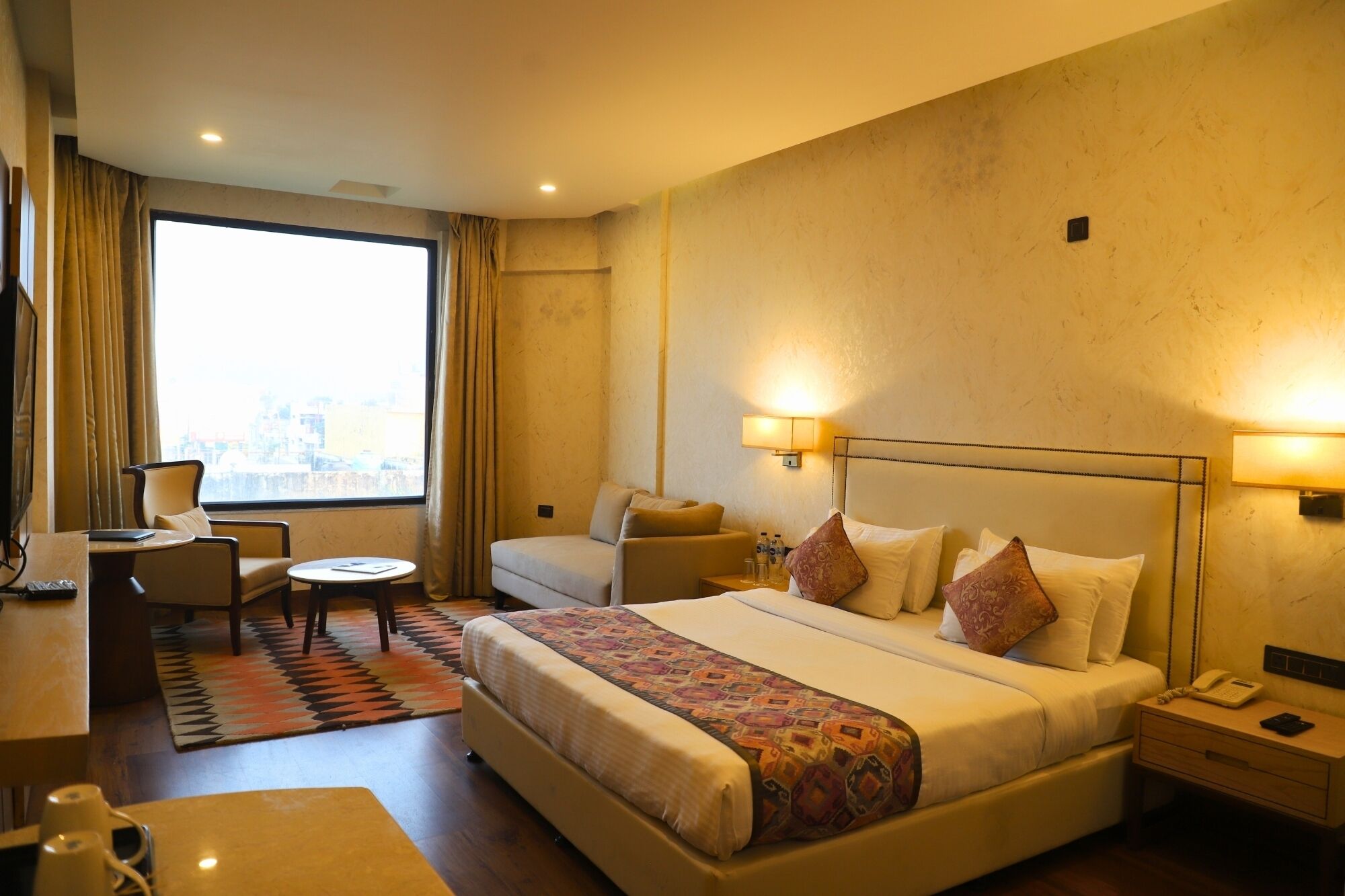 A hotel room with a large bed, two bedside tables, a desk with a chair, a small round table, and a large window with curtains.