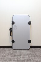 Une valise fermée, dotée d’une poignée et de roulettes, est posée contre un mur, sur un sol recouvert d’une moquette.