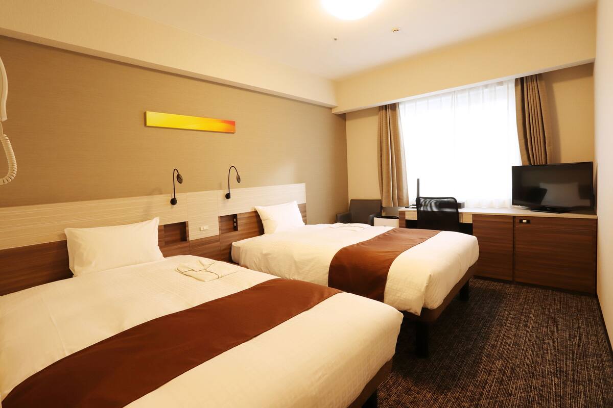 Ein Hotelzimmer mit zwei Betten, einem Schreibtisch, einem Fernseher, einem Telefon und einem Fenster mit Vorhängen.