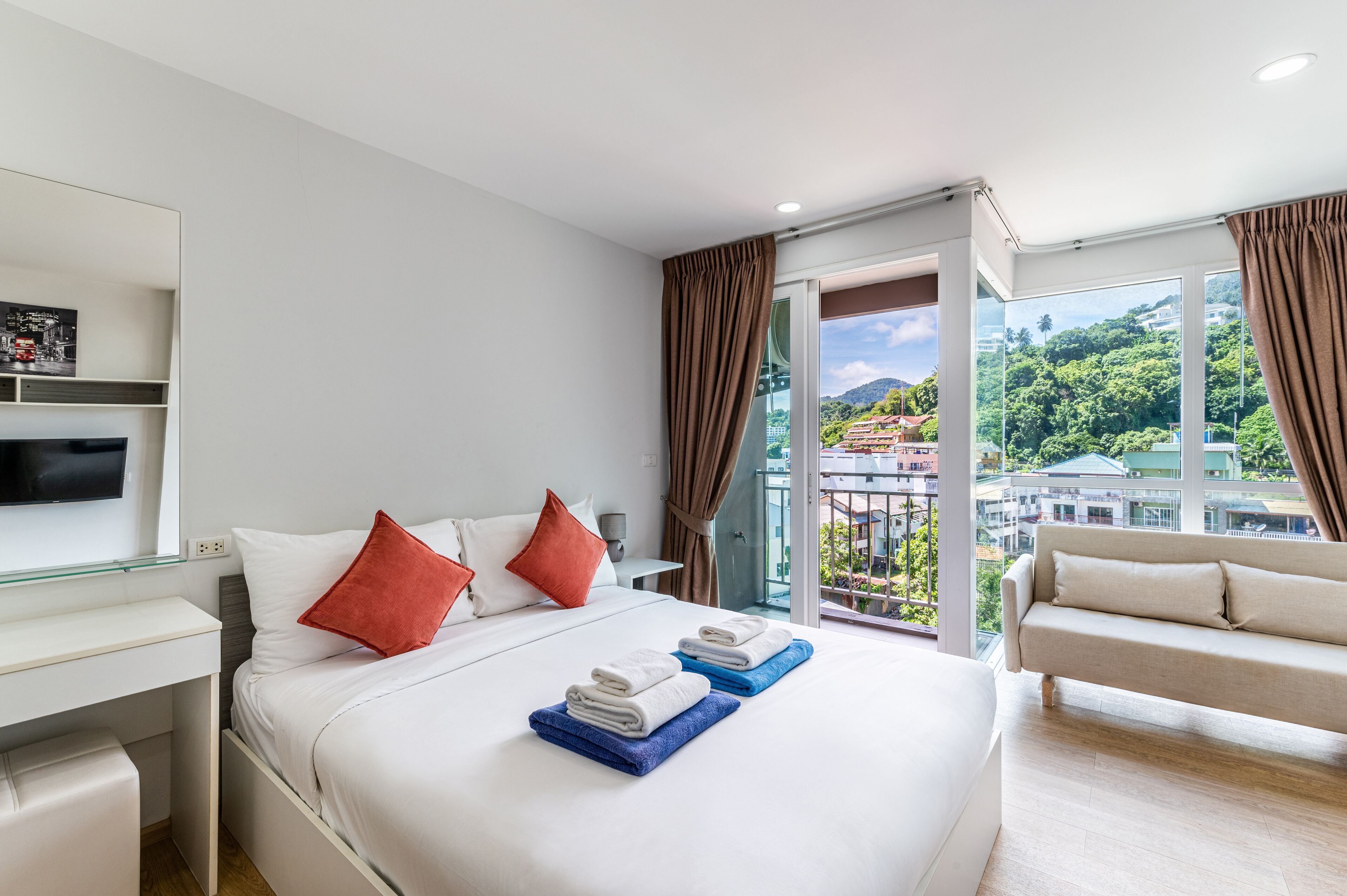 A modern hotel room with a large bed, a desk, a TV, and a balcony with a city view.