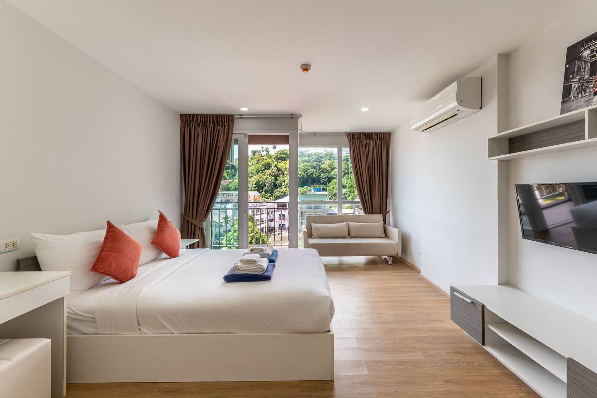 A modern hotel room with a large bed, a sofa, a TV, and a balcony with a view of buildings and trees.