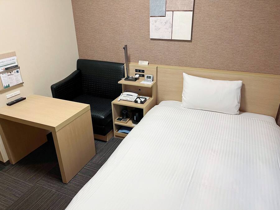 A hotel room with a bed, a desk, a chair, a nightstand with a phone and a clock, and a bulletin board with a notice.