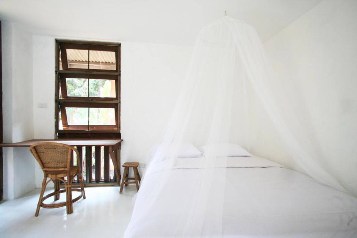 Ein Zimmer mit einem Bett, das von einem weißen Moskitonetz verdeckt ist, einem Holztisch mit einem Stuhl und einem kleinen Hocker.
