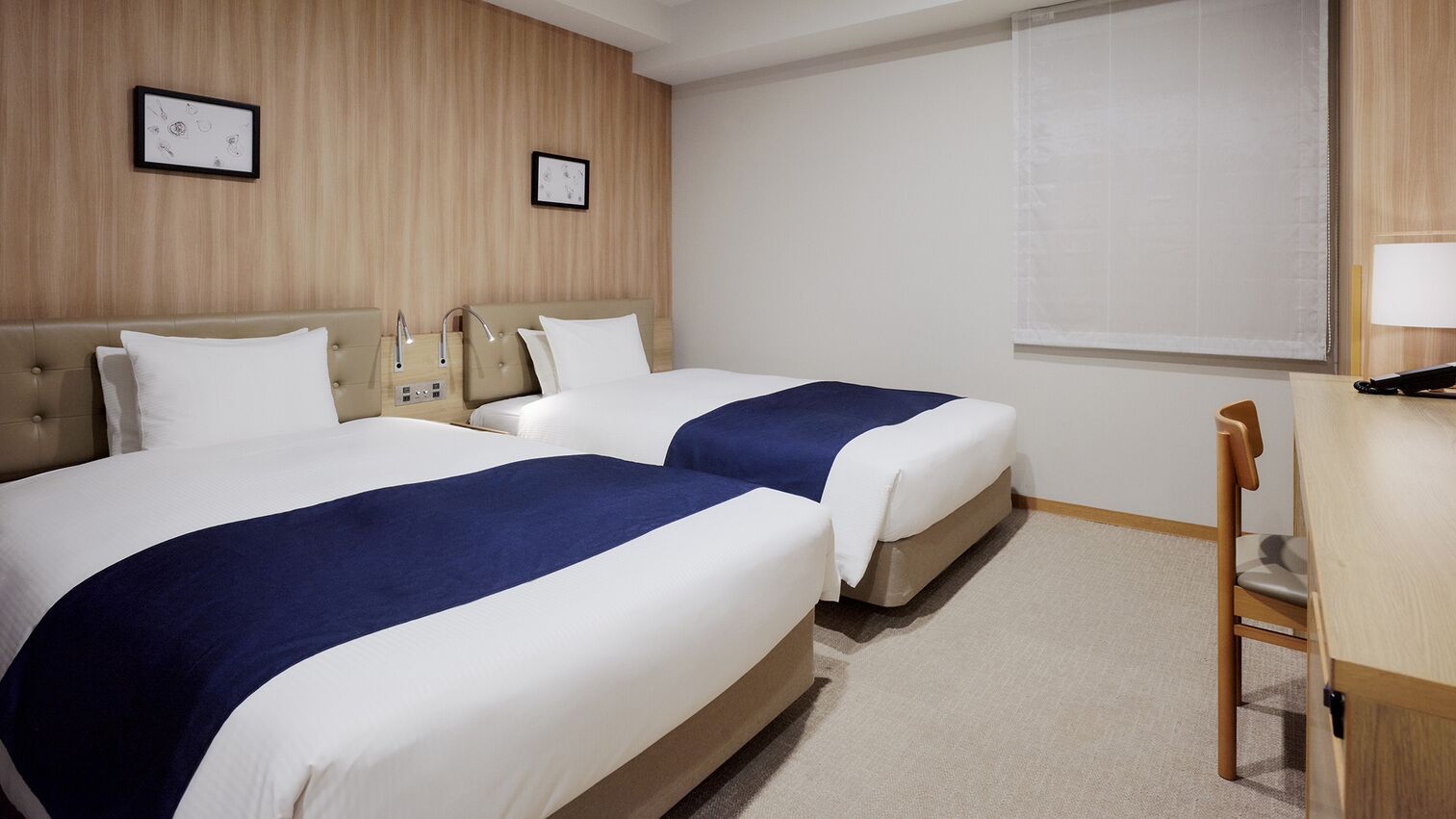 A hotel room with two beds, a wooden headboard, a bedside table, a lamp, and a window with blinds.