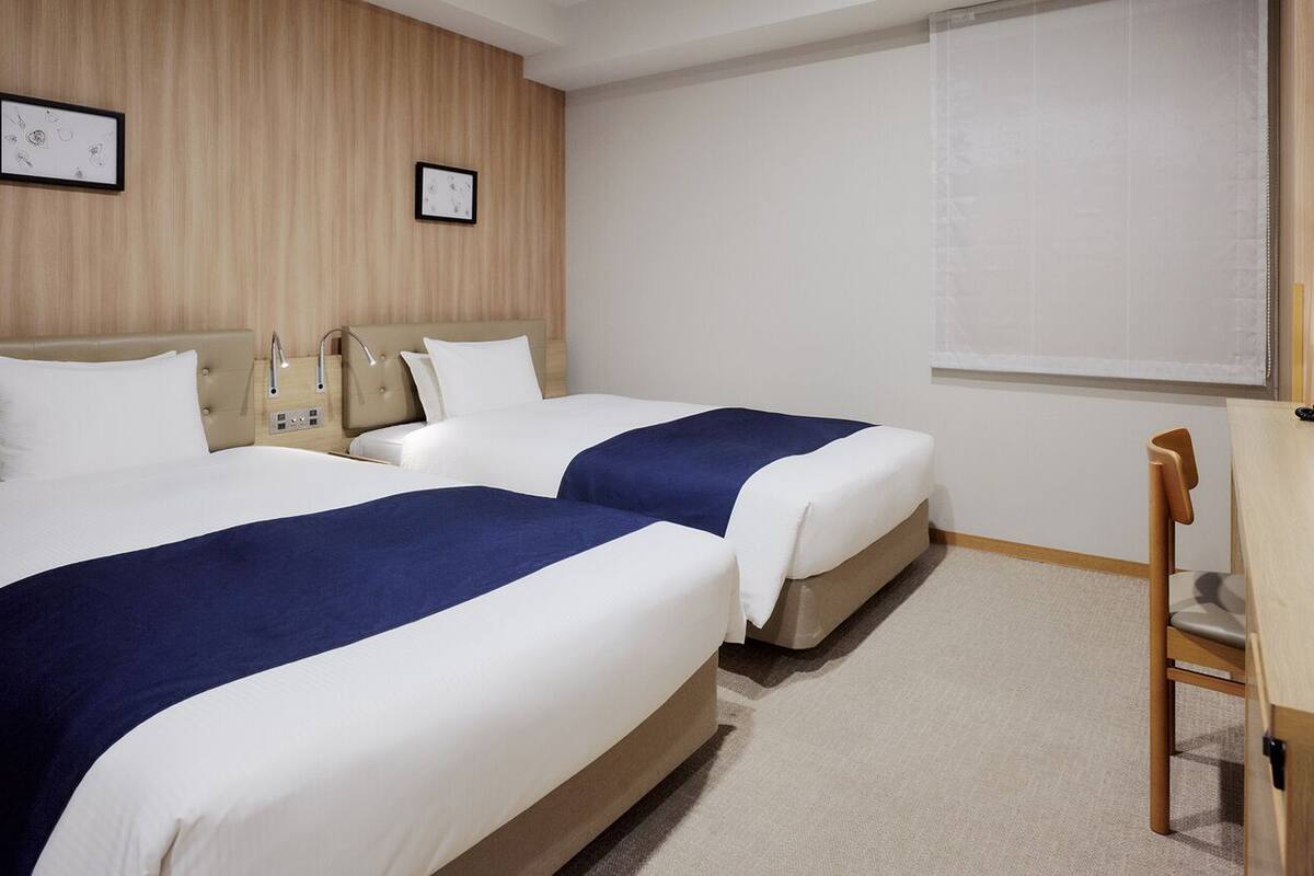 A hotel room with two beds, a wooden headboard, a bedside table, a lamp, and a window with blinds.