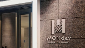 L’extérieur de l’hôtel MONday HANEDA AIRPORT se distingue par son design moderne, avec une entrée en verre et un logo bien visible sur le mur.