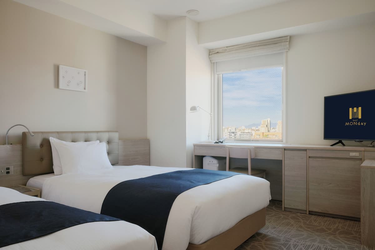 A hotel room with two beds, a desk, a TV, and a window with a city view.