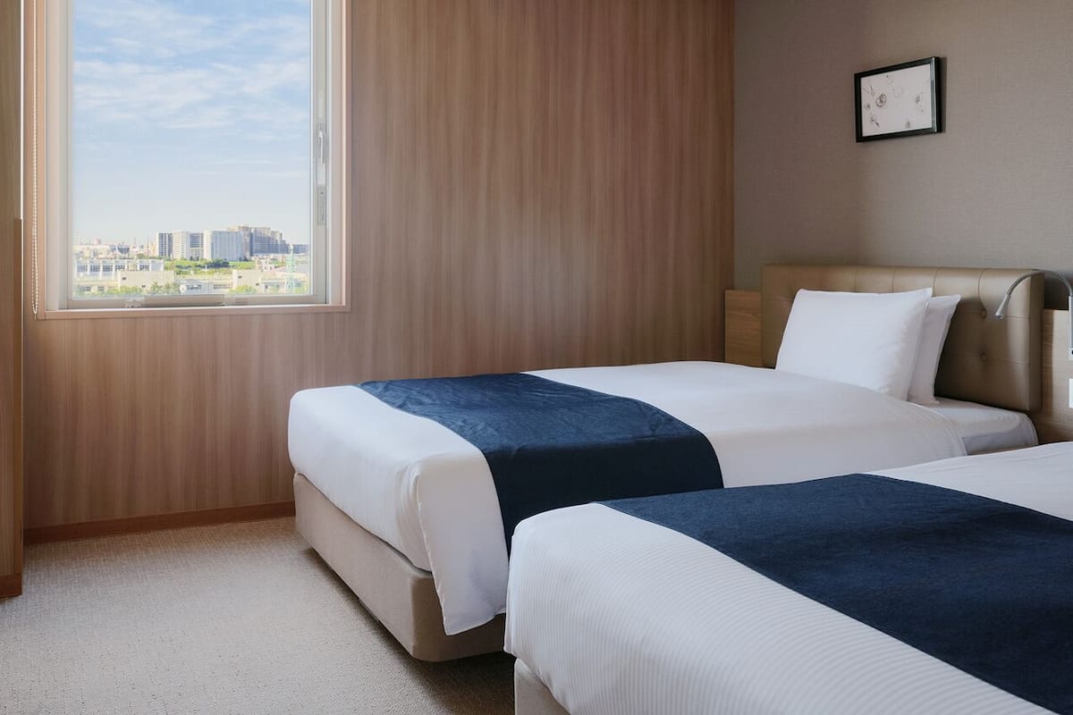 A hotel room with two beds, a desk, a chair, and a large window with a city view.