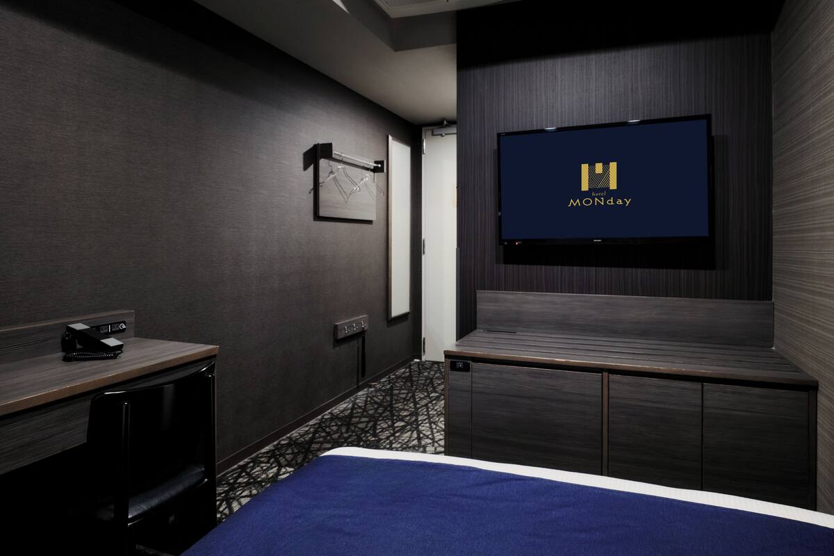 A hotel room with a bed, a TV mounted on the wall, a desk with a chair, and a coat rack.