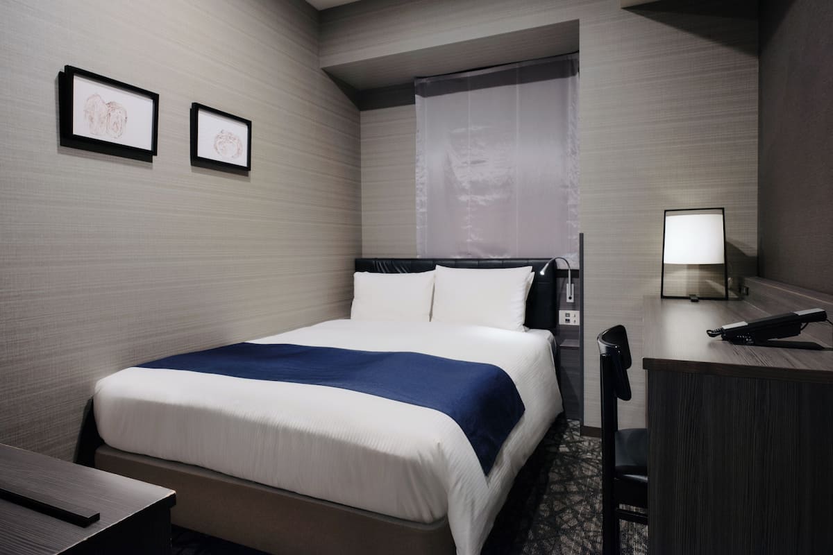 A hotel room with a bed, a desk, a chair, a lamp, and two framed pictures on the wall.