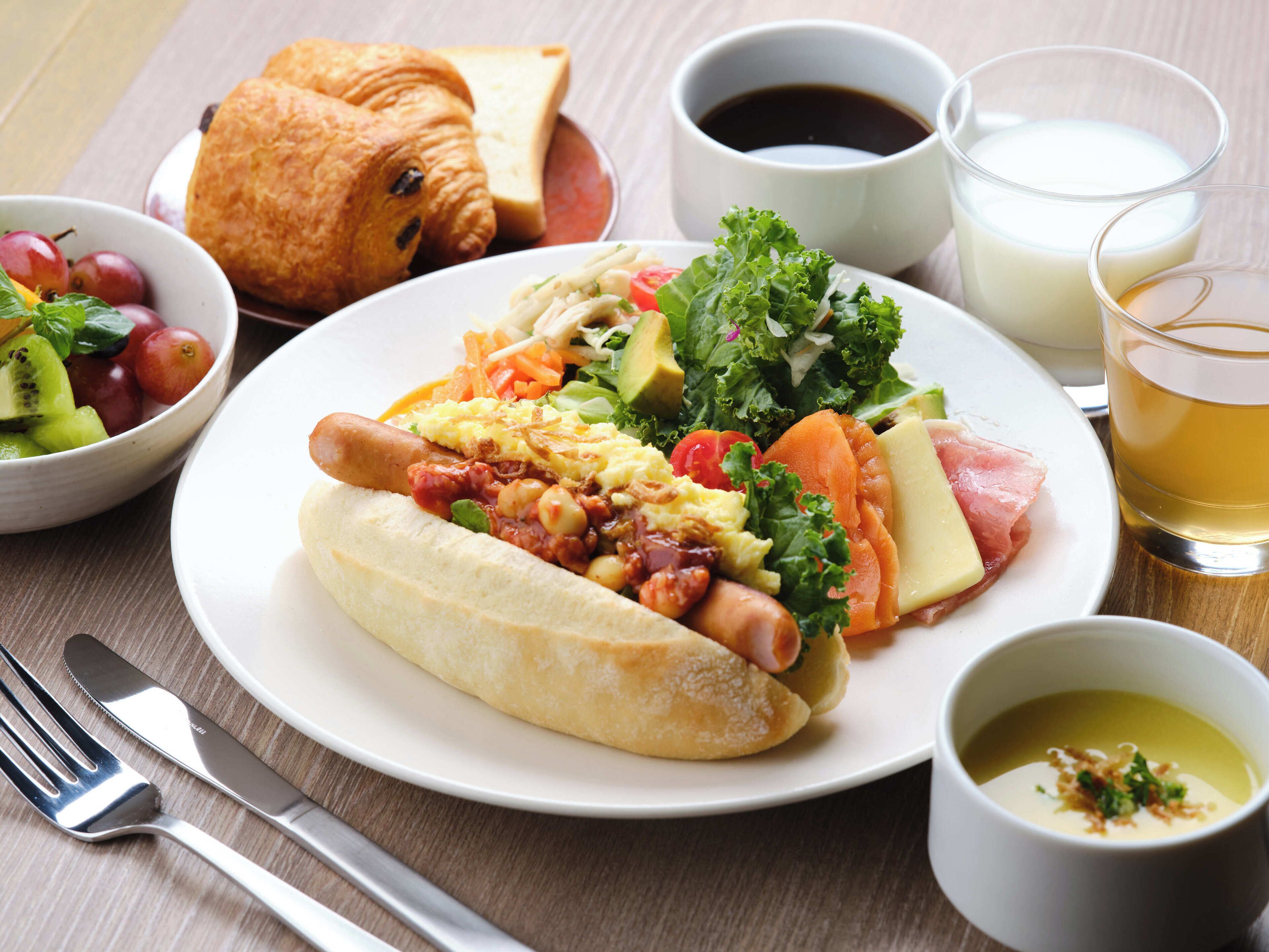 Um prato com um cachorro-quente, vegetais e um croissant. Há também potes com frutas, uma xícara de café, um copo de leite e uma xícara de chá.