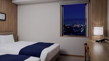 Een hotelkamer met twee bedden, een bureau, een lamp en een raam met uitzicht op de stad.
