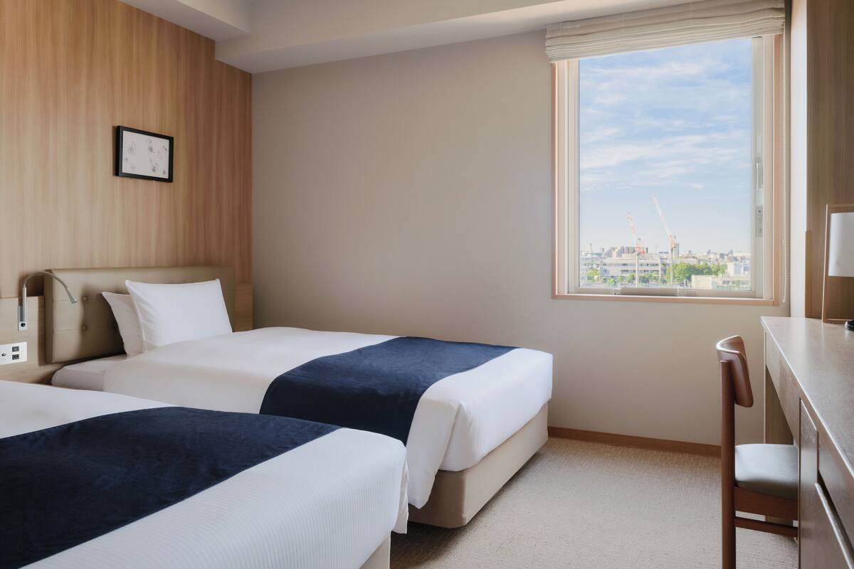 A hotel room with two beds, a desk, and a window with a city view.