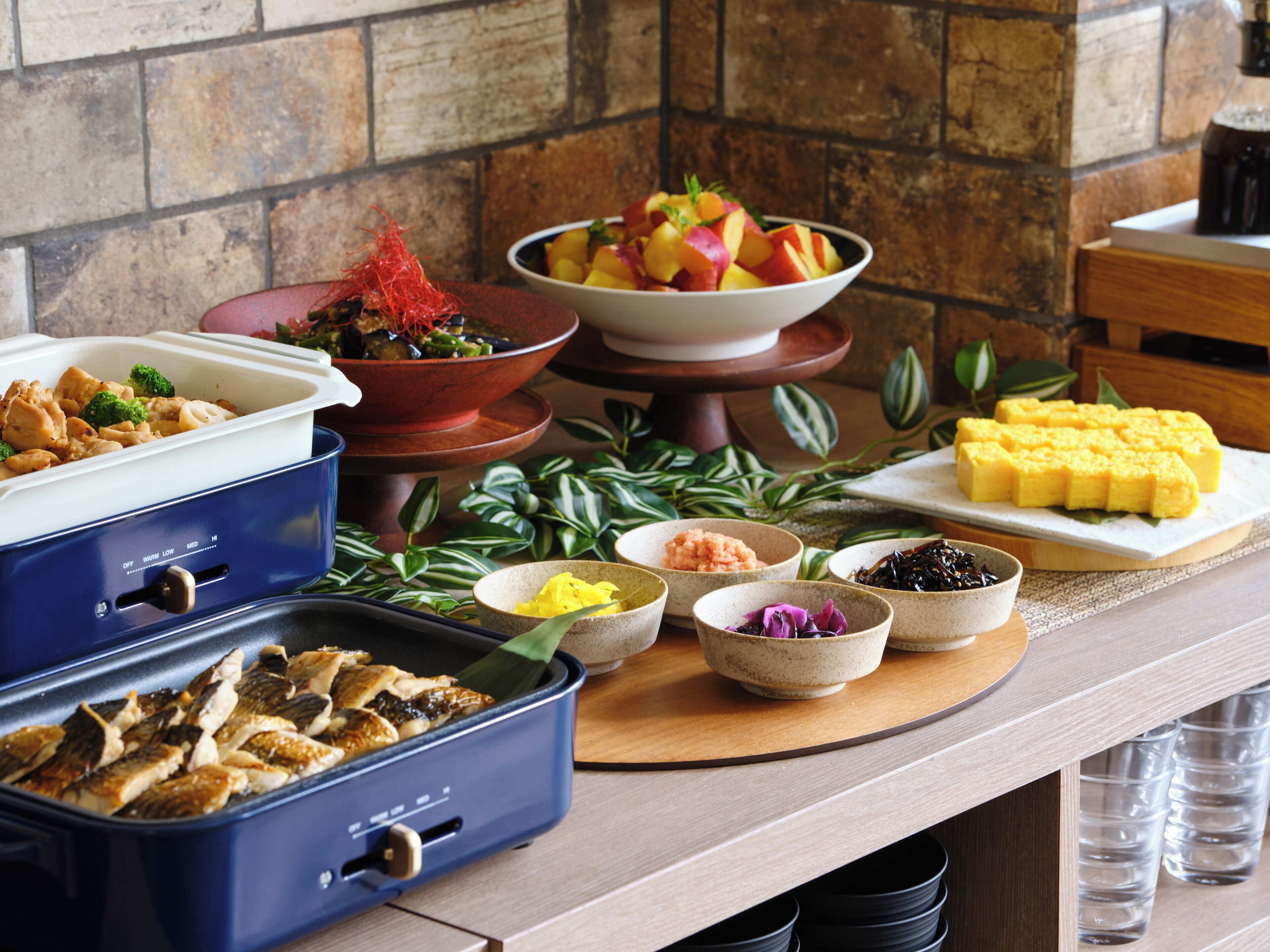 A buffet table with various dishes, including a bowl of fruit salad, a dish with broccoli, and a container of fish.