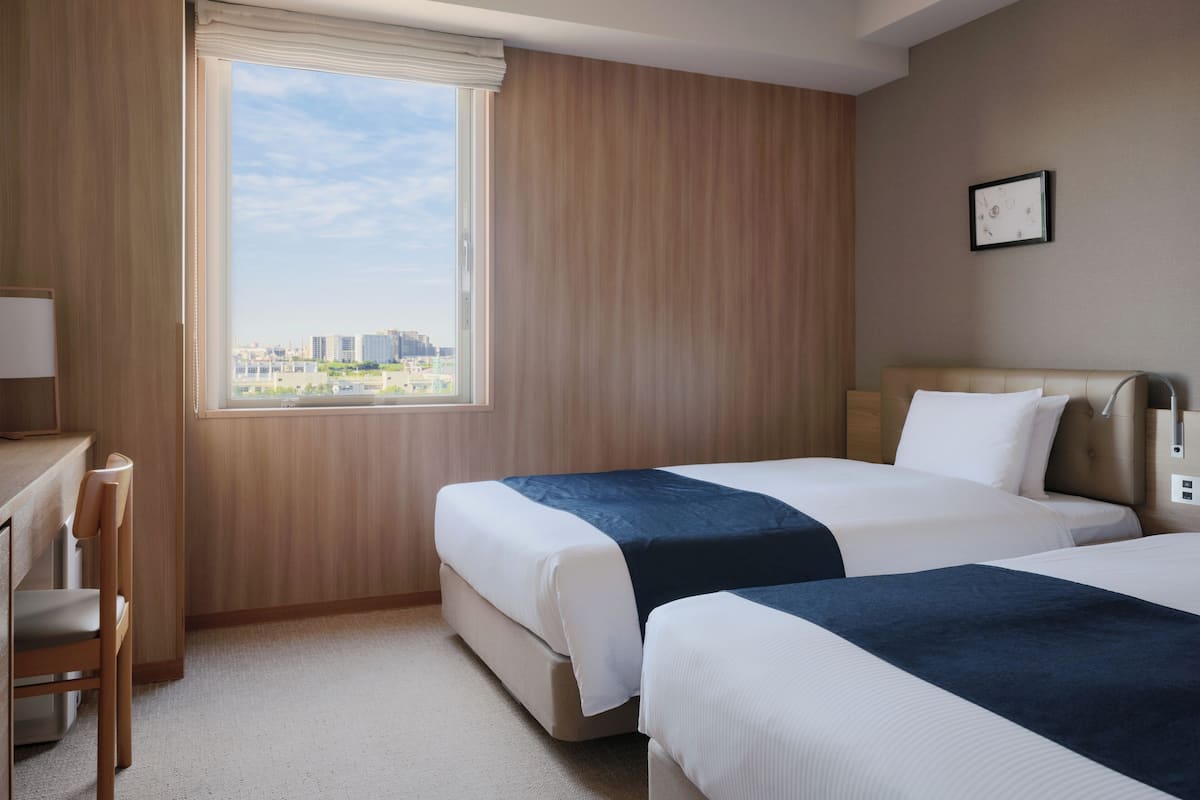 A hotel room with two beds, a desk, and a window with a city view.