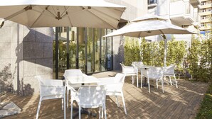 An outdoor seating area with white chairs and large umbrellas, a wooden deck, and a building with glass windows in the background. - Hotel MONday Haneda Airport (Tokyo)