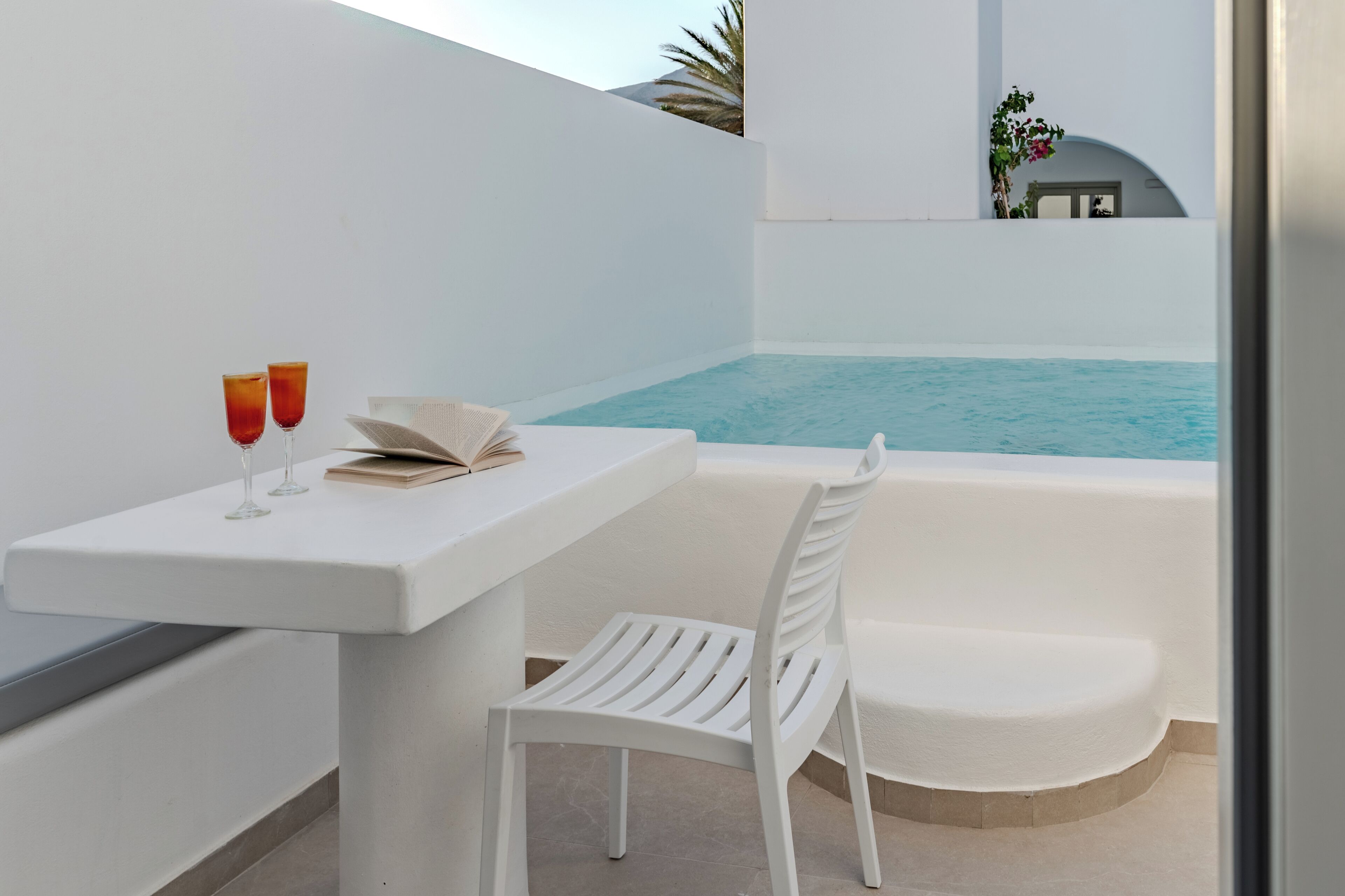 A white balcony with a pool, a white table with two glasses and a book, and a white chair.