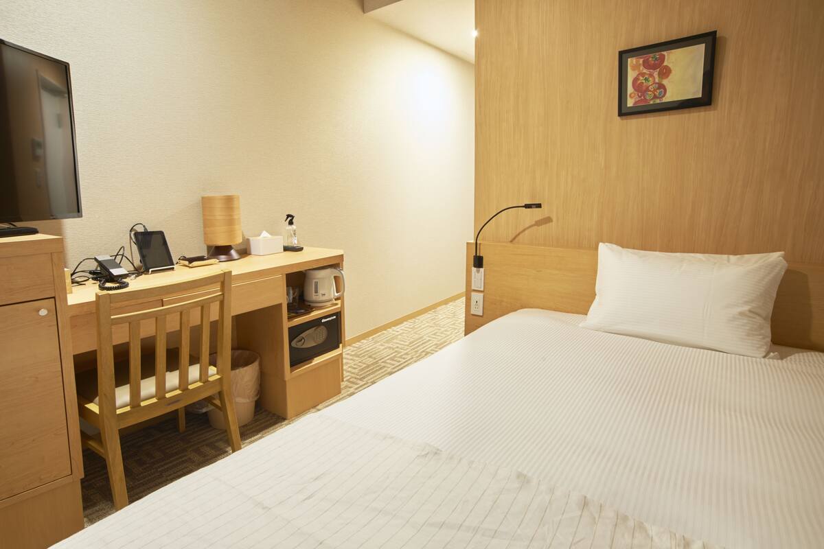 A hotel room with a bed, a desk with a chair, a television, and a small table with a lamp and a tissue box.