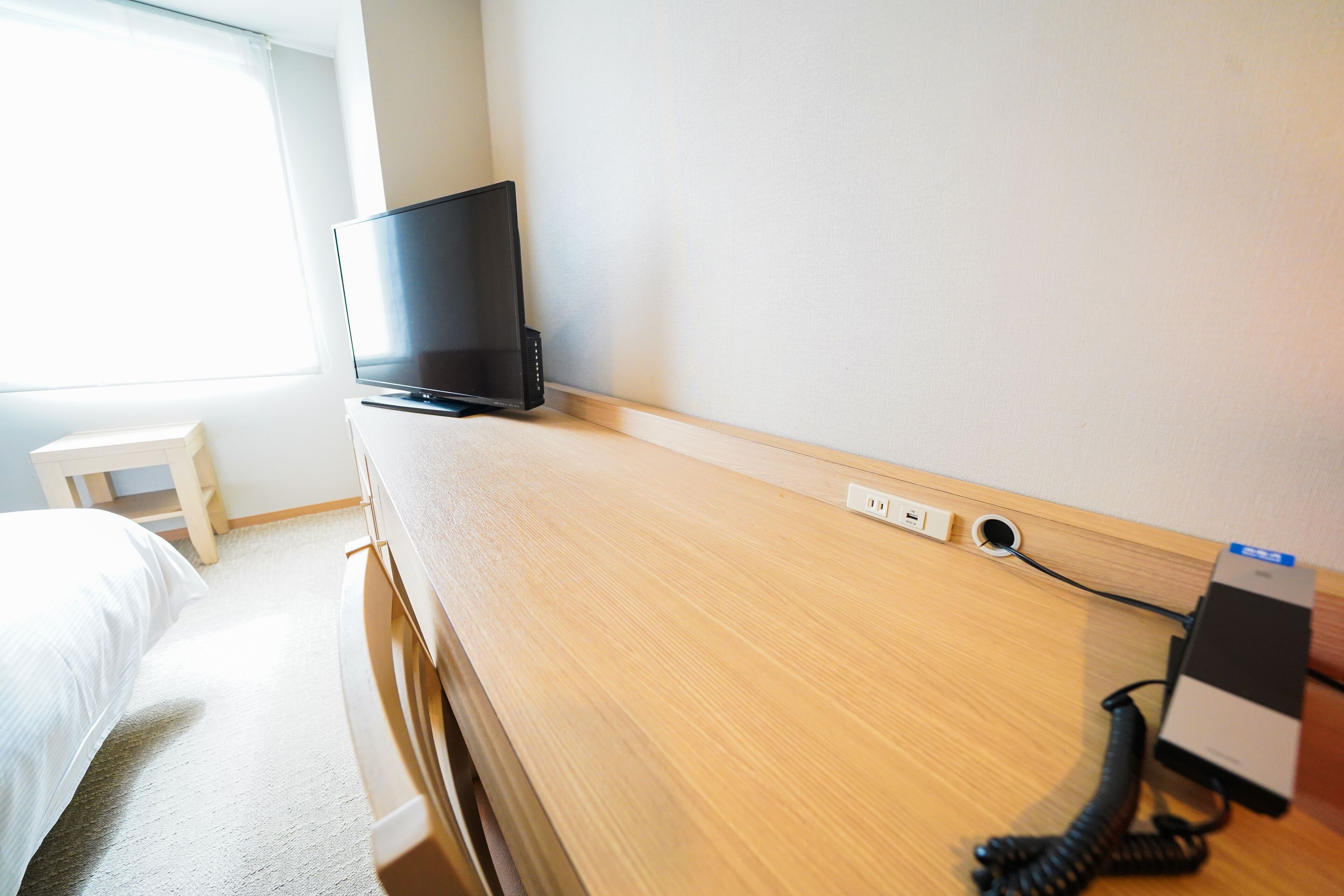A hotel room with a bed, a TV on a wooden stand, a desk with a phone, and a window.