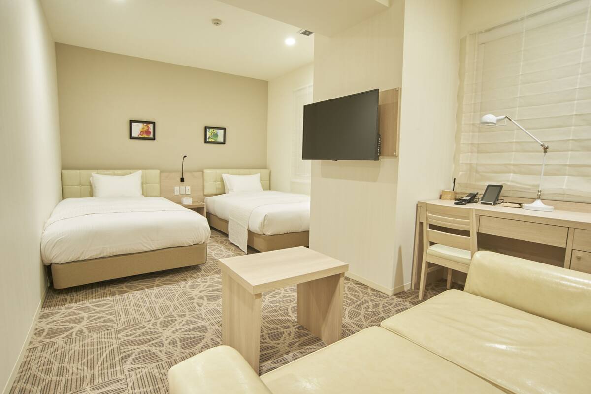 A hotel room with two beds, a desk, a chair, a TV, and a lamp.