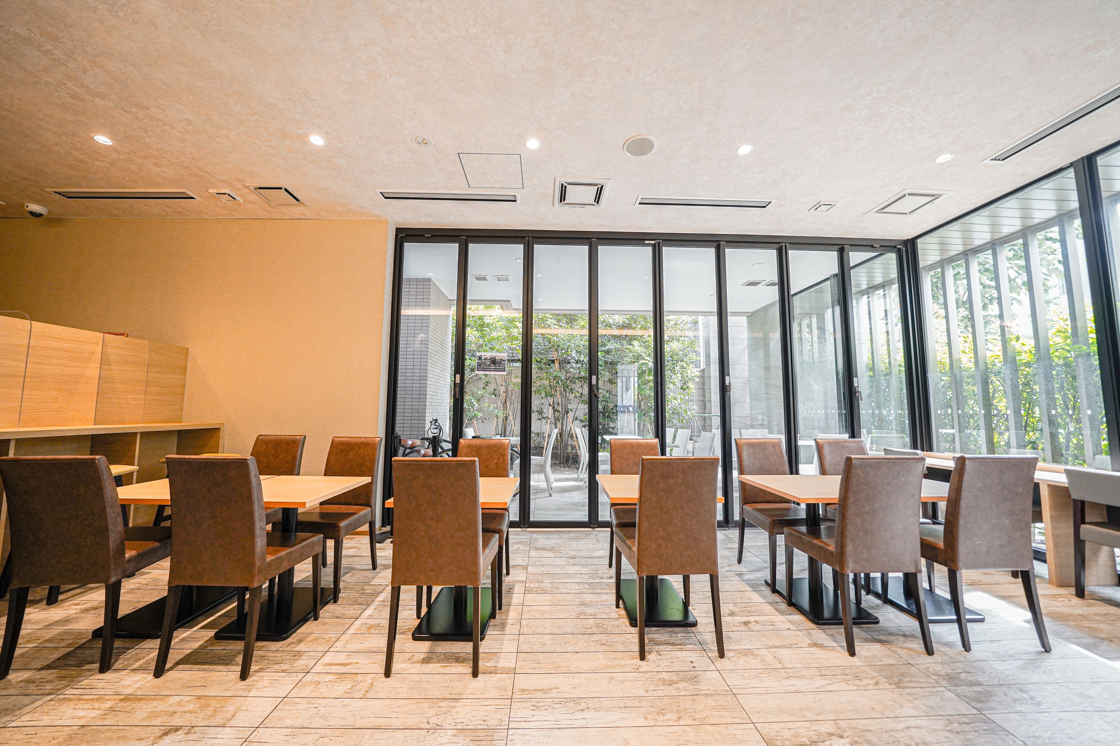 A modern dining area with multiple tables and chairs, large windows, and a view of greenery outside.