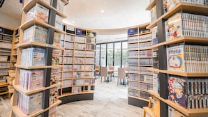 A bookstore with curved wooden shelves filled with manga and comics. There are chairs and tables in the center for reading. - Quintessa Hotel Tokyo Haneda Comic & Books (Tokyo)